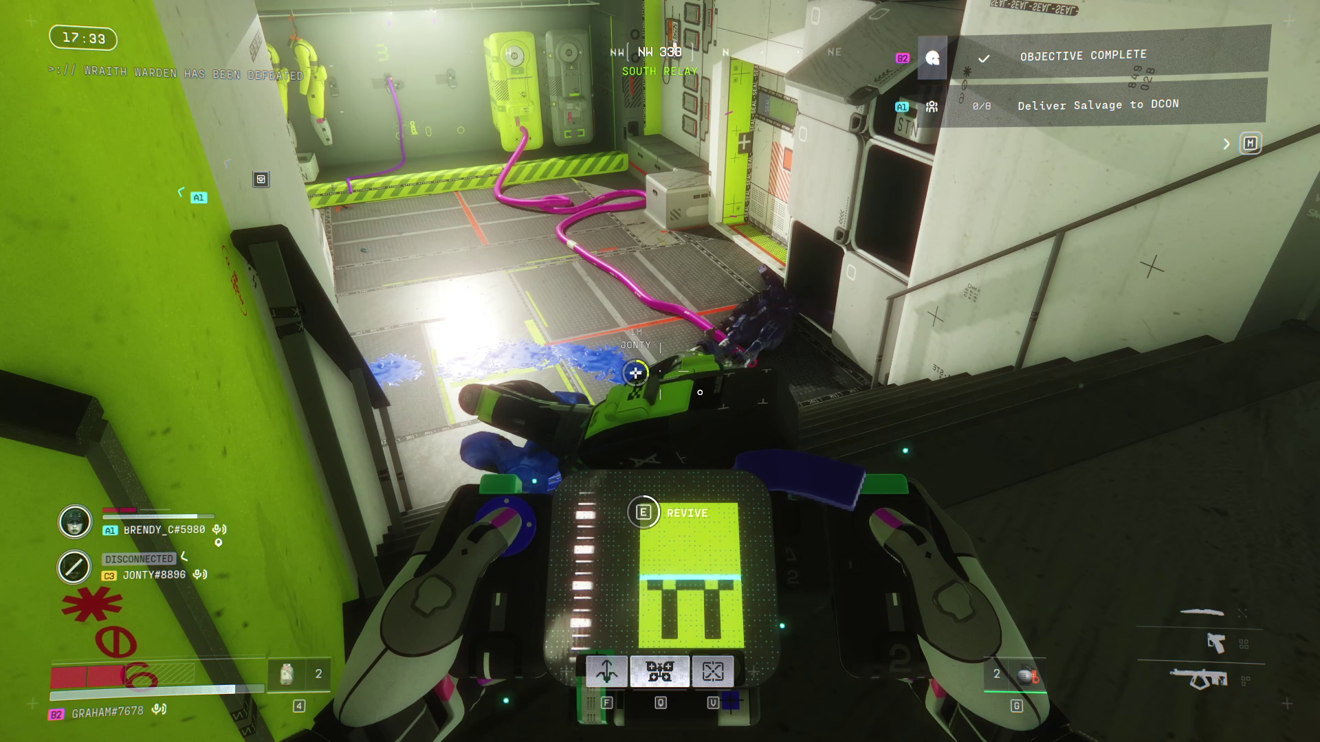In a green stairwell, the player holds a device up to another player who is lying on the ground. The device says 'Revive' on it.